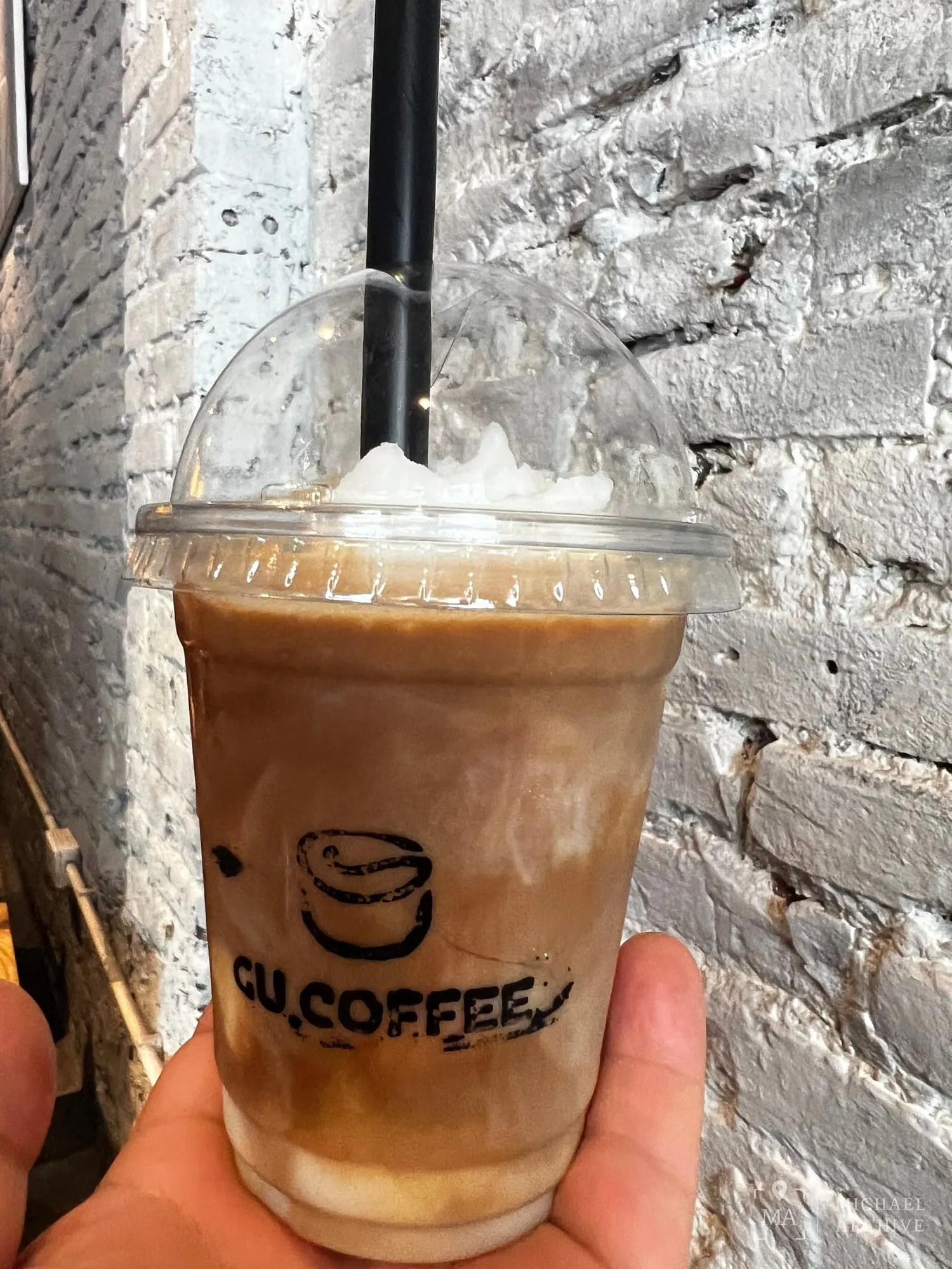 흰 벽돌 벽 앞에서 손에 든 GU.COFFEE 아이스커피