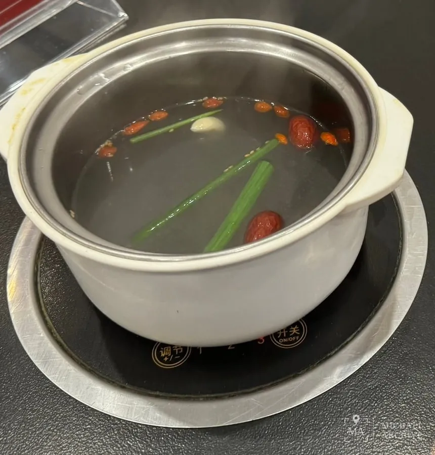 흰 뚝배기에 맑은 육수가 담겨 있다. 대추와 구기자, 마늘, 쪽파가 떠 있고 테이블에 박힌 인덕션 위에 올라가 있다.