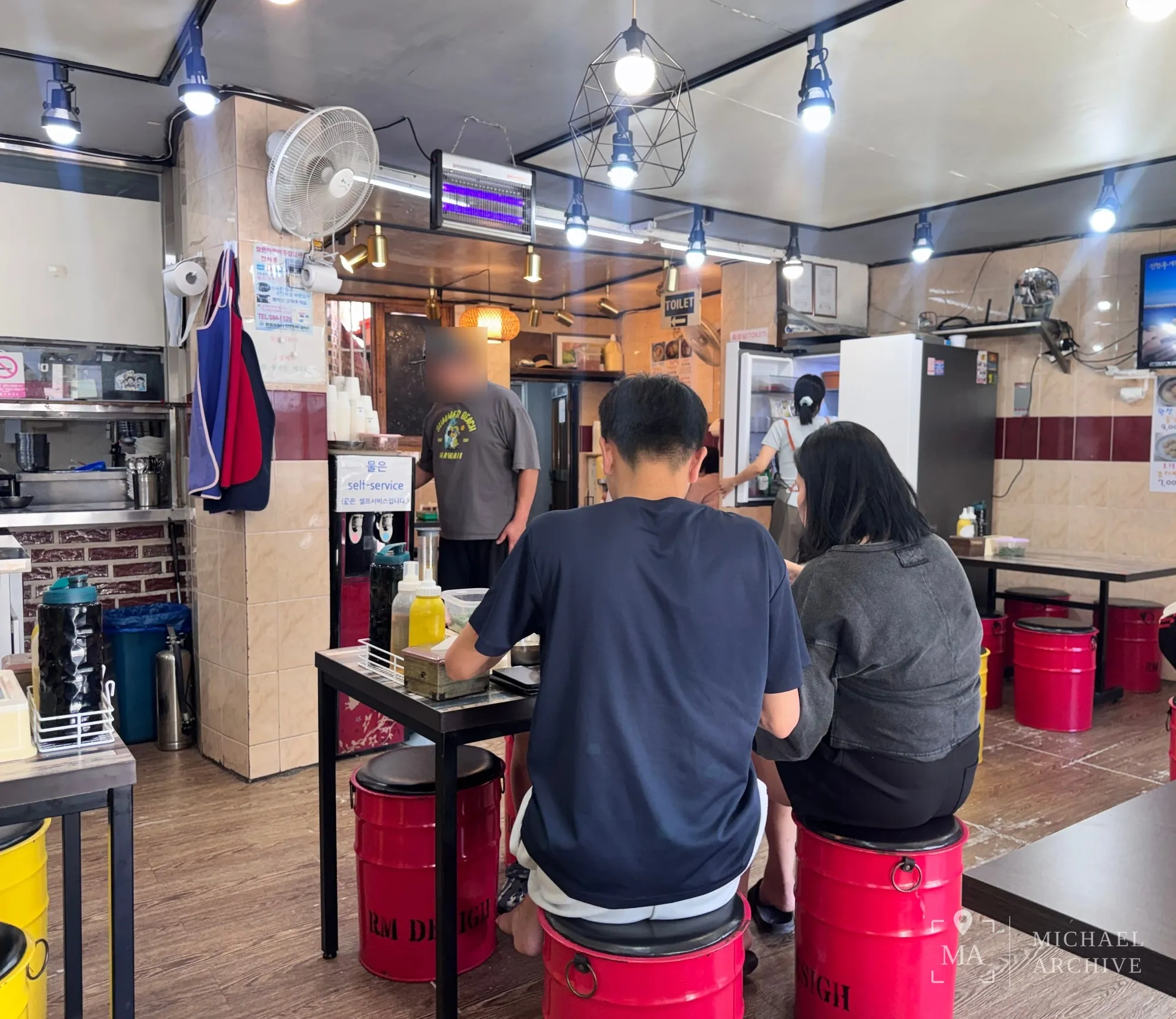 매장 안 — 빨간 드럼통 의자, 천장 선풍기, ‘물은 self-service’ 안내