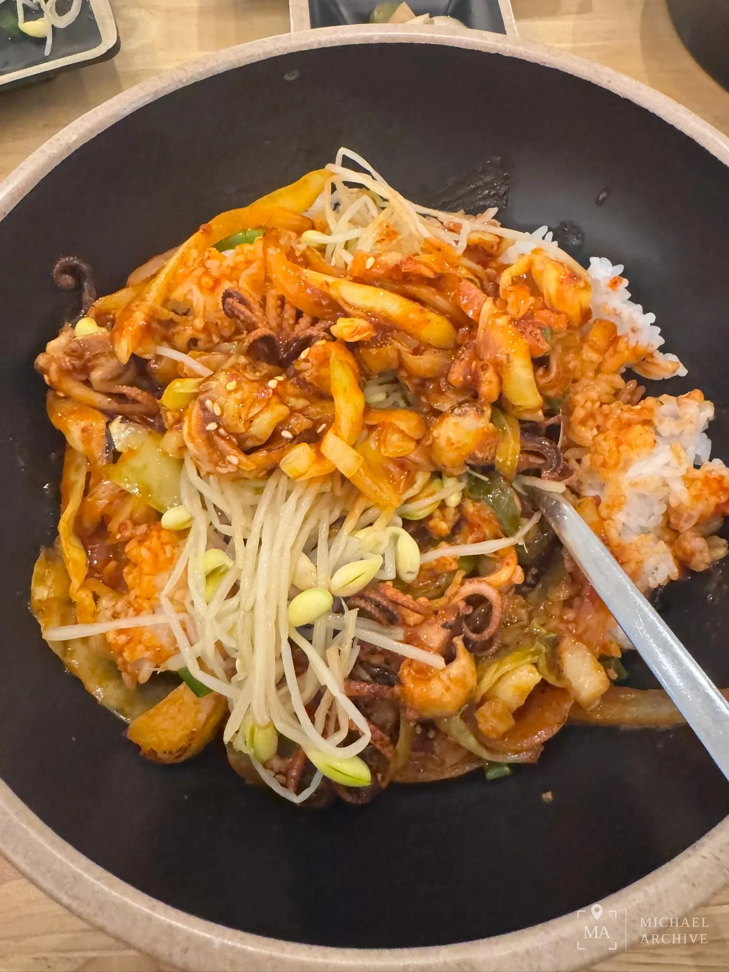 낙지볶음 클로즈업. 붉은 양념에 낙지 다리, 양배추, 애호박, 양파, 콩나물이 섞여 있고 오른쪽 위로 흰밥이 보인다