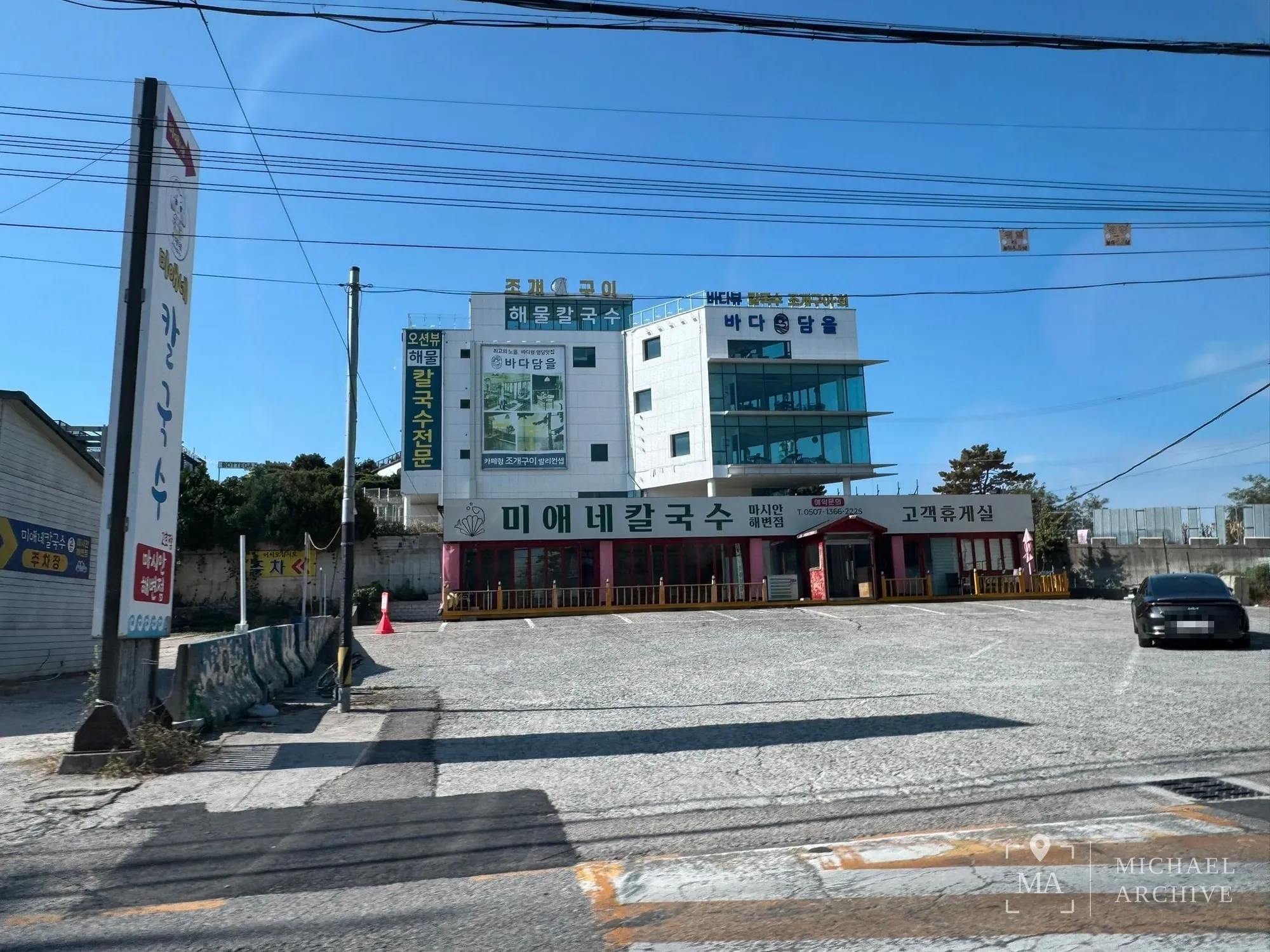 넓은 자갈 주차장 너머로 「미애네칼국수 마시안해변점 · 고객휴게실」 간판이 붙은 낮은 별채가 길게 서 있다. 왼쪽에 「주차장」 안내판