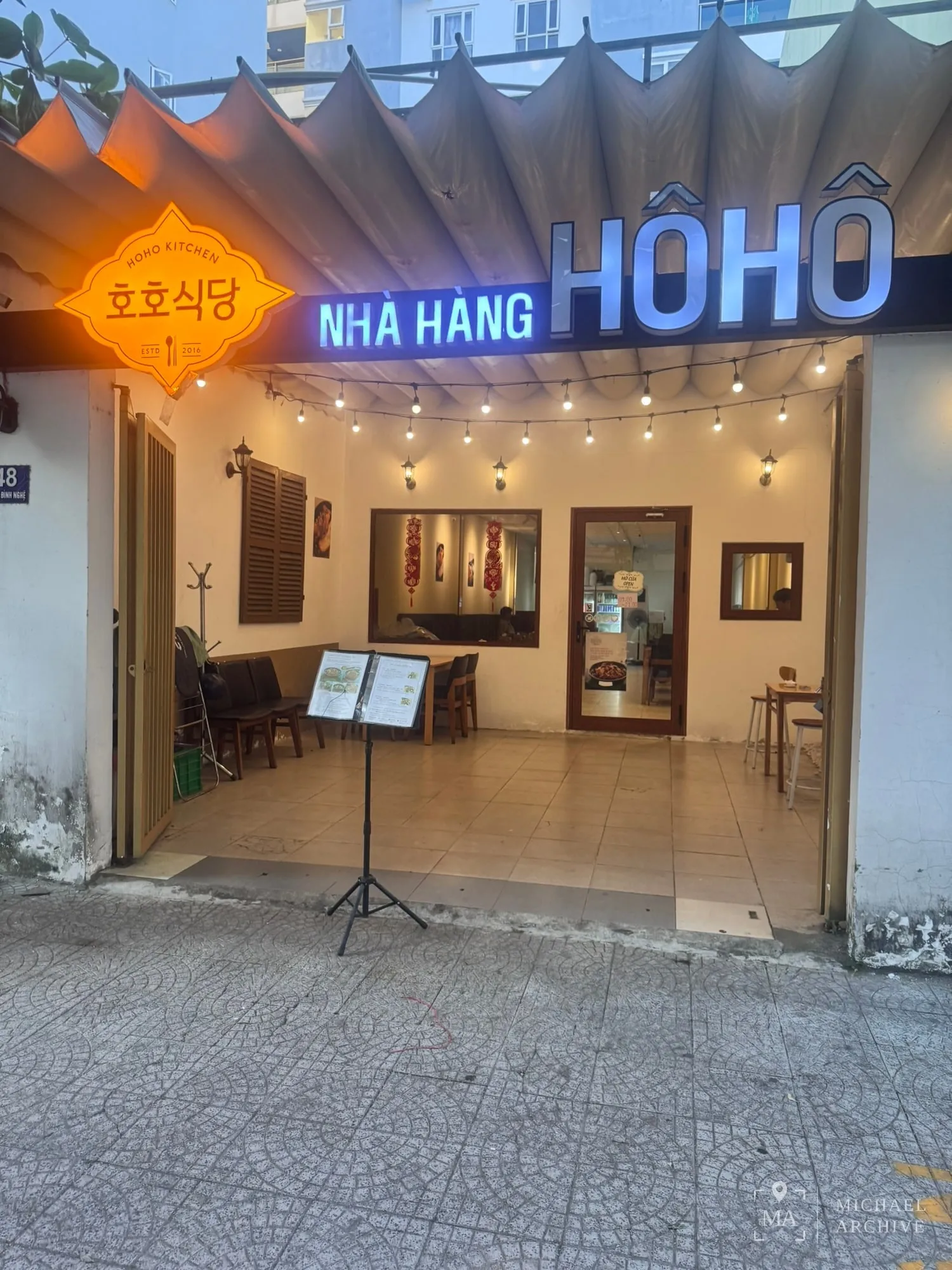 해가 진 직후, 호호식당 입구. 왼쪽에 「호호식당 HOHO KITCHEN ESTD 2016」 주황색 간판, 오른쪽에 「NHÀ HÀNG HÔHÔ」 파란 네온 간판이 걸려 있다
